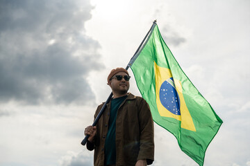 Portrait of cheerful young man looking forward while standing proudly with holding Brasil flag over...