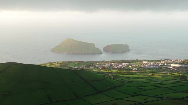 Aerial view of Pico Dona Joana, where vibrant green fields meet the ocean, contrasting with the distant islands under a soft, hazy sky, geod&eacute;sico, Portugal.
