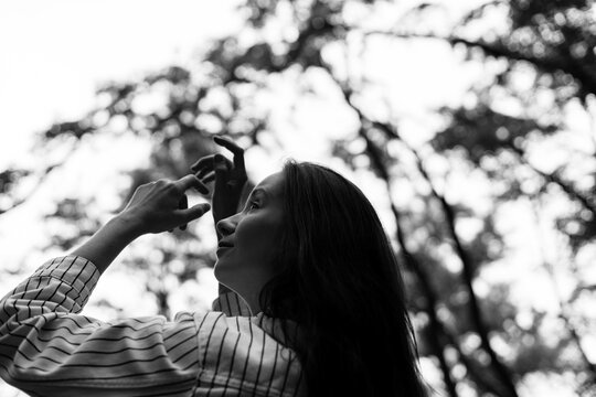 A woman reaches up to the sky with arms raised over the head, looking up. A black and white photo of a girl back view, with a forest on background. People meditating or simply relaxing alone in nature - Powered by Adobe