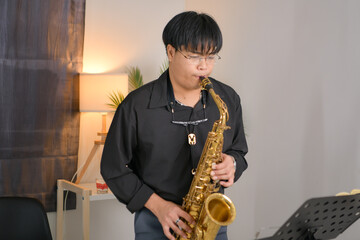 Young Asian man playing saxophone indoors in a cozy room with soft lighting