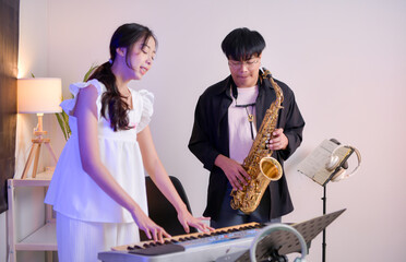 Two young Asian man and woman performing music indoors with keyboard and saxophone