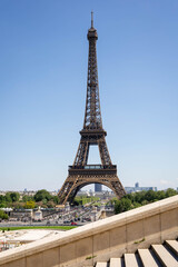 Obraz premium Low-angle view of the Eiffel Tower on a bright, sunny day, showcasing its intricate metalwork against a clear blue sky, with Parisian cityscape and crowds visible in the background.