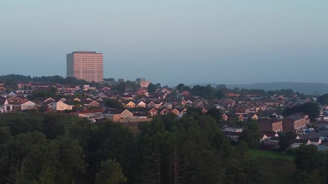 Sideways Travelling Drone Shot of Houses Surrounding Large Office Building with Trees in Foreground with Soft Sunrise Light. Drone View of Densely Populated Estates and Streets.