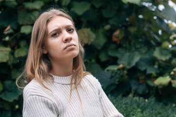 Naklejka premium A young woman with long hair sits outside, displaying a thoughtful and serious expression. Lush greenery in the background enhances her contemplative mood during daylight.