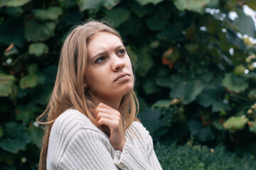 A woman with long hair contemplates while sitting outdoors surrounded by vibrant green foliage. The...