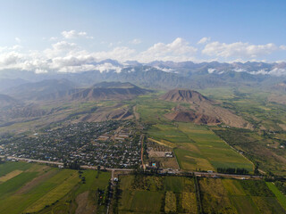 A wide aerial shot of a small town nestled between green fields and majestic mountains under a cloudy sky
