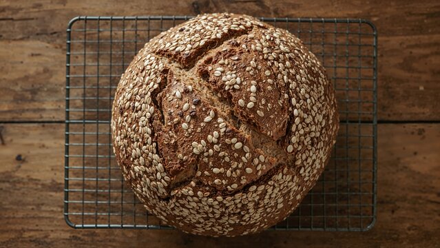 Flat lay of a loaf of whole grain bread with seeds cooling on a wire rack atop a wooden table