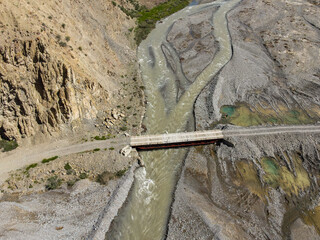 A top-down aerial view of a simple bridge crossing a muddy, rocky river in a remote, arid canyon landscape