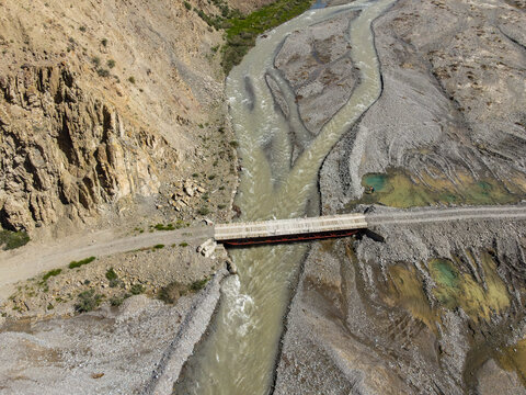 A top-down aerial view of a simple bridge crossing a muddy, rocky river in a remote, arid canyon landscape