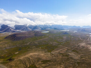 A wide aerial view of a remote mountain landscape with scattered lakes, wetlands, a dirt road, and snow-capped peaks in the distance