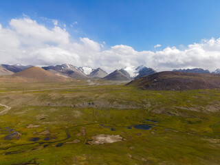 A wide aerial view of a remote mountain landscape with green fields, small ponds, and snow-capped peaks in the far distance under a bright sky