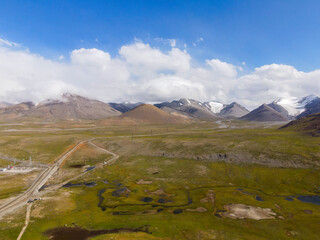 An aerial view of a rugged, remote mountain landscape with green and brown fields, wetlands, and snow-capped peaks in the distance