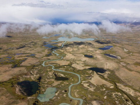 A wide aerial view of a winding river and small lakes in a remote, rugged mountain landscape under a cloudy sky with distant peaks