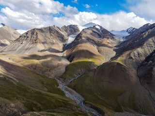 A drone shot of a dramatic mountain landscape with rugged peaks, glaciers, and a river flowing through a valley under a cloudy sky