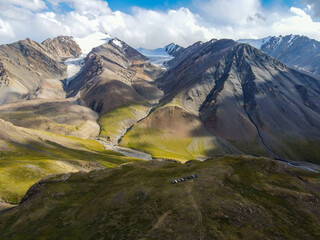 A drone shot of a group of vehicles parked on a small hill in a dramatic, rugged mountain landscape with snow-capped peaks