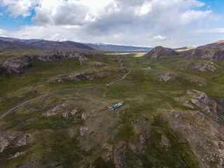 An aerial view of a line of cars parked on a dirt path in a rugged, remote mountain landscape with scattered rocks, green grass, and small lakes