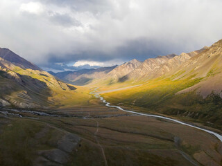 A drone shot of a river winding through a wide mountain valley with dramatic light, stormy clouds, and rugged, textured slopes