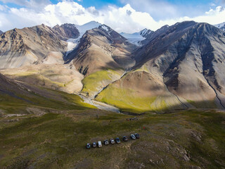 An aerial view of a group of cars and people parked on a hill in a dramatic, rugged mountain landscape with snow-capped peaks and glaciers