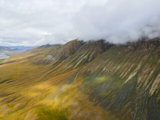 A drone shot of a vast mountain slope with green and yellow tones, with clouds rolling over the peak in a remote, wild landscape