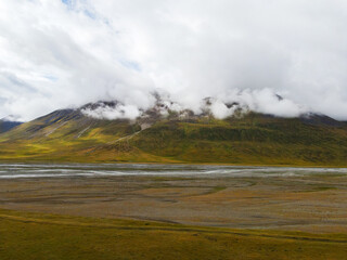 A wide aerial view of a rugged mountain with clouds rolling over the peak, overlooking a vast, flat plain with a braided river