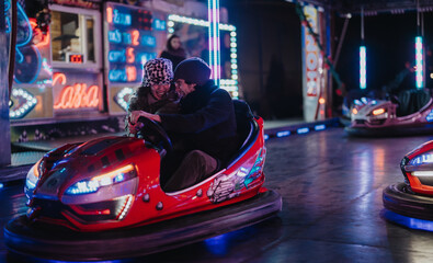 A cheerful couple riding bumper cars at an evening amusement park surrounded by colorful lights....