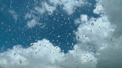 Condensation and drops of rain or dew on a transparent vehicle windshield with a blue sky and clouds behind. Illustrates concepts of storm, rainfall, spring, cloudscape, and typhoon.