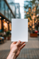 Hand holding vertical blank flyer on festive shopping street with warm bokeh lights. Perfect for branding and holiday promotions. Copy space included.