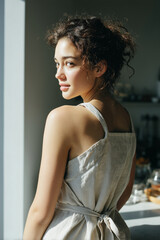 Young woman in natural linen apron standing in warm sunlight. Calm domestic lifestyle scene with soft natural light. Copy space included.