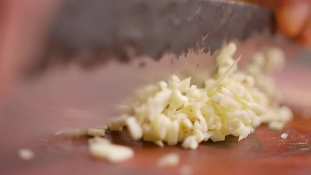 Close up of a chefs hands using a sharp kitchen knife to mince fresh garlic cloves into small pieces on a wooden cutting board 