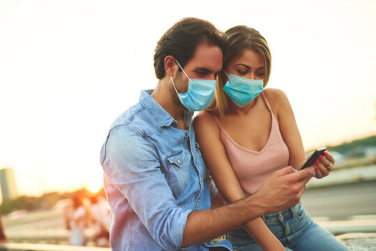 Couple enjoying sunset while wearing masks and using smartphone in urban setting