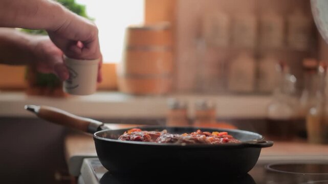 A man's hands sprinkle spices into a hot, steaming pan where an Osso Buco steak is simmering. Spices and herbs are blurred in the background. Gourmet home cooking in progress