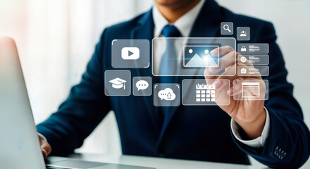 A businessman in a suit and tie interacts with a futuristic digital interface displaying various icons for business, technology, and communication, all while seated at a desk with a laptop