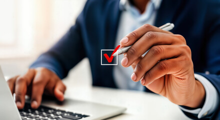 A businessmans hand holding a pen, marking a checkmark in a box on a document while working on a laptop in an office setting, symbolizing task completion and decisionmaking