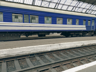 A view of a blue passenger car standing on a platform under a glass station roof. The railway track is visible in the foreground, symbolizing movement and new journeys.