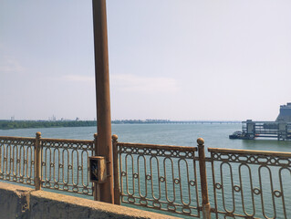 A majestic river under an endless sky, with a metal railing on the bridge in the foreground. The distant bank with greenery and elements of urban development is visible.
