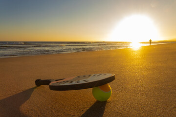 Beach tennis racket and ball lying on golden sand during a colorful sunset. Perfect scene for summer outdoor sports and seaside recreation.