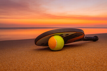Peaceful sunset sky reflecting on the ocean with beach tennis racket and ball in the foreground. Represents outdoor activity and tropical seaside mood.