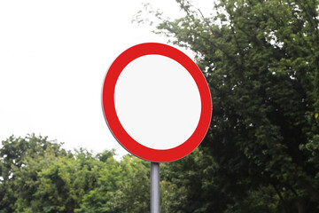 Blank traffic sign with red border. Circular road symbol with no text. Roadside pole with empty warning sign. Mockup for transport message. Green trees in background. Daylight outdoor setting.