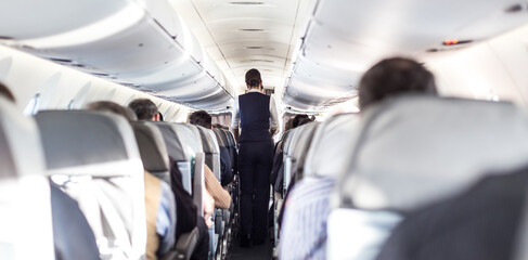 Interior of airplane with passengers on seats and stewardess in uniform walking the aisle, serving...