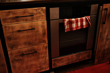 Close-up of a rustic wooden kitchen cabinet and a modern built-in oven with a red checkered towel...