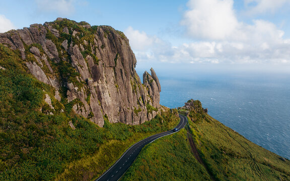 Aerial view of jagged cliffs juxtapose with the serene ocean, while a winding road traces the contours of the verdant landscape, Rocha dos Frades, Azores, Portugal.