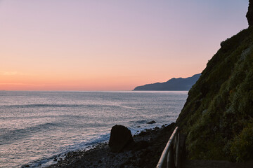 Serene coastal landscape at twilight, featuring a rocky beach, calm ocean waves, and a lush green...