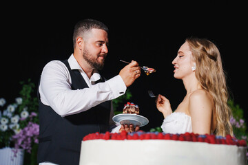 Bride and groom eat a large round cake decorated with berries in the night.