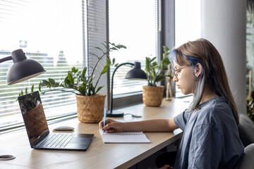 Focused female student sitting at desk with laptop, taking notes and studying online. Concept of...