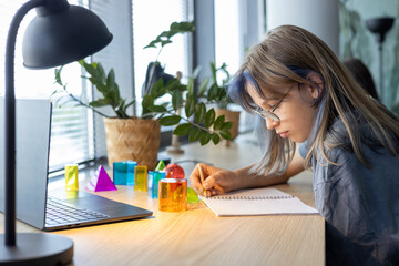 Teen girl studying 3D geometric shapes during math class, drawing figures in notebook and exploring spatial concepts through hands-on learning and interactive school project.