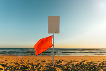 Red warning flag on a sandy beach with ocean waves in the background. Symbol of dangerous swimming conditions and beach safety under sunny weather with clear blue sky.