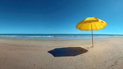 A bright yellow beach umbrella casts a shadow on the sand under a clear blue sky by the ocean.