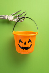 Bright vertical orange pumpkin-shaped bucket in Halloween style hanging from a skeleton hand © ActionGP