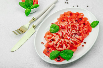 Sliced Coeur de Boeuf tomato with balsamic drops and basil leaves on an oval plate. Vegan-friendly