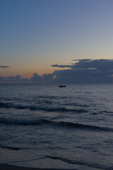 Fototapeta premium A small boat floats alone on the dark ocean at dusk, with the sky displaying dramatic clouds and hues of orange and blue.